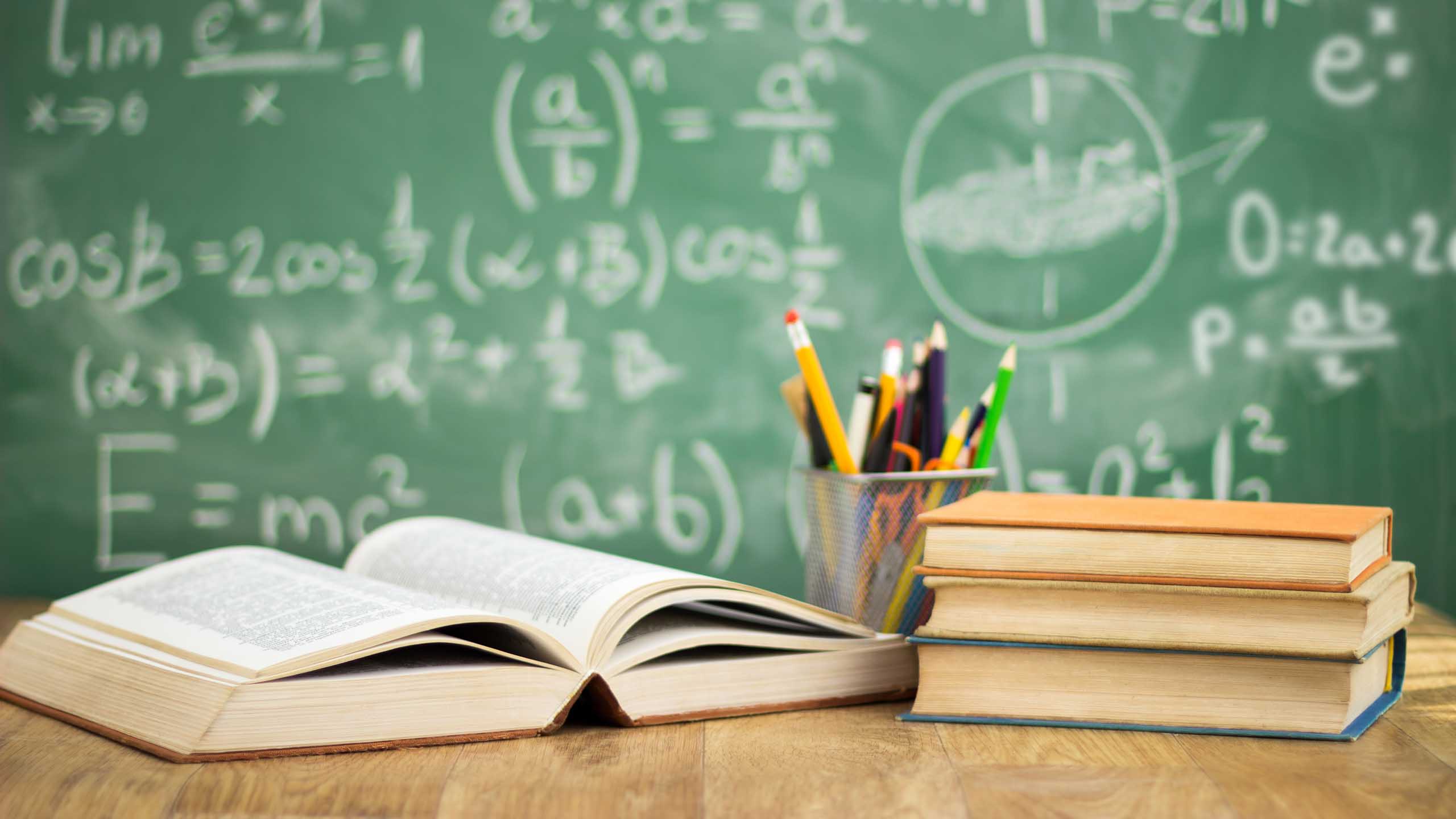 Skolböcker och pennor på ett skrivbord framför en skrivtavla med matematiska formler.